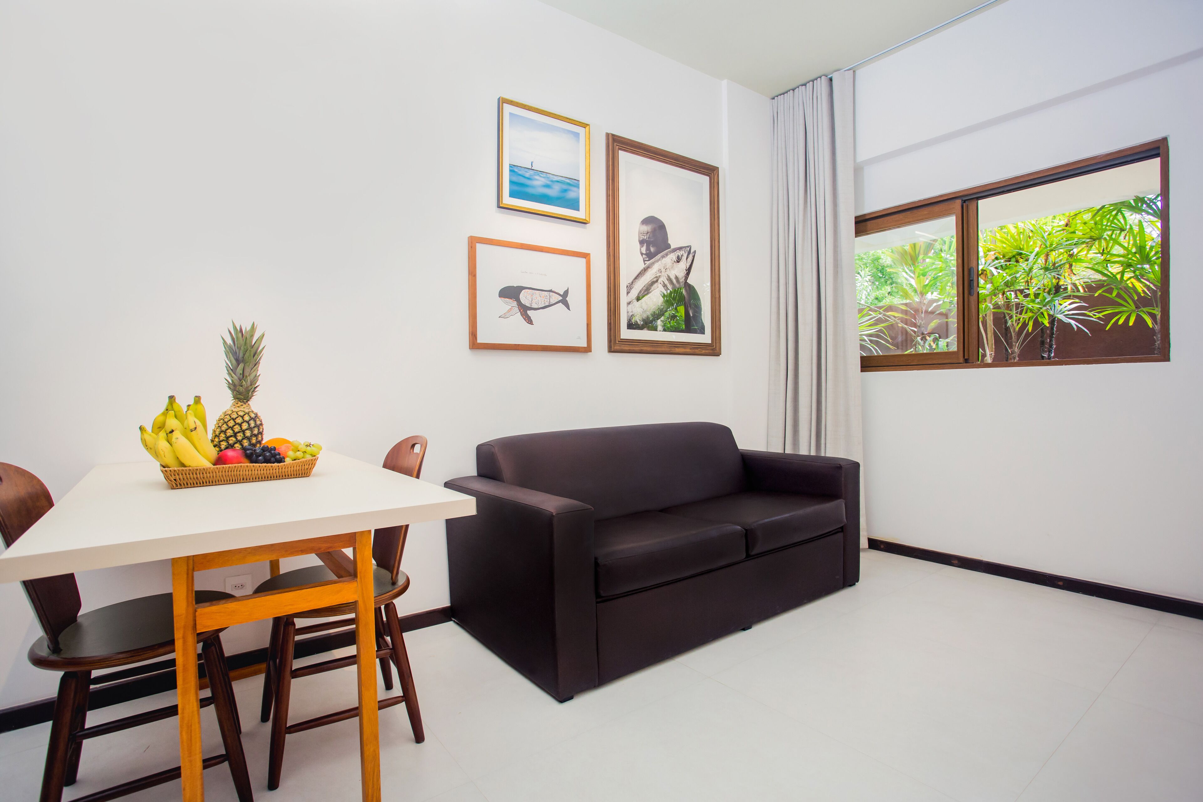 A modern living room with a black sofa, a dining table with chairs, and a fruit basket. There are framed pictures on the wall and a window with a view of greenery.