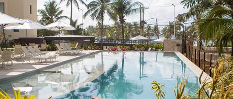 Un área de piscina con sillones de descanso y sombrillas, rodeada de palmeras.