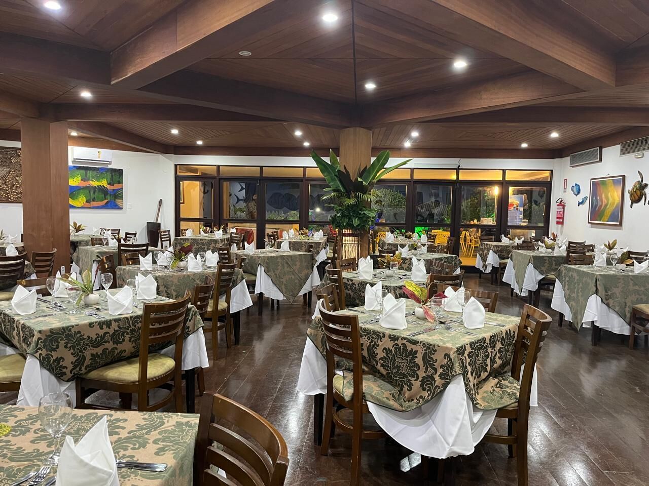 A restaurant with multiple tables covered with patterned tablecloths, wooden chairs, and a central plant.