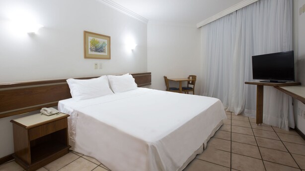 Una habitación de hotel con una cama grande, una mesita de noche con teléfono, una silla, un escritorio con televisor y un cuadro en la pared.