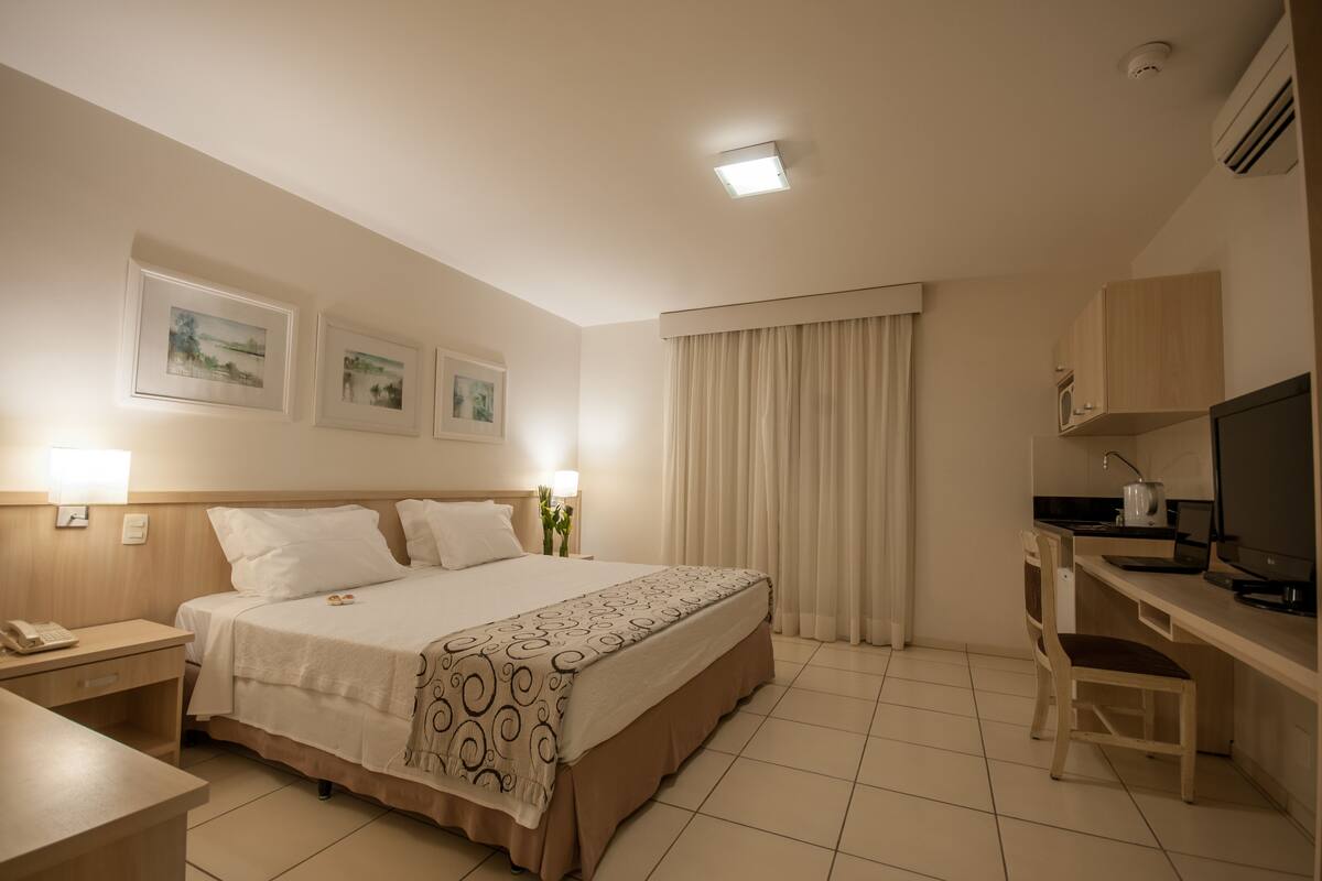 A hotel room with a bed, bedside table, lamp, desk, chair, TV, and curtains.