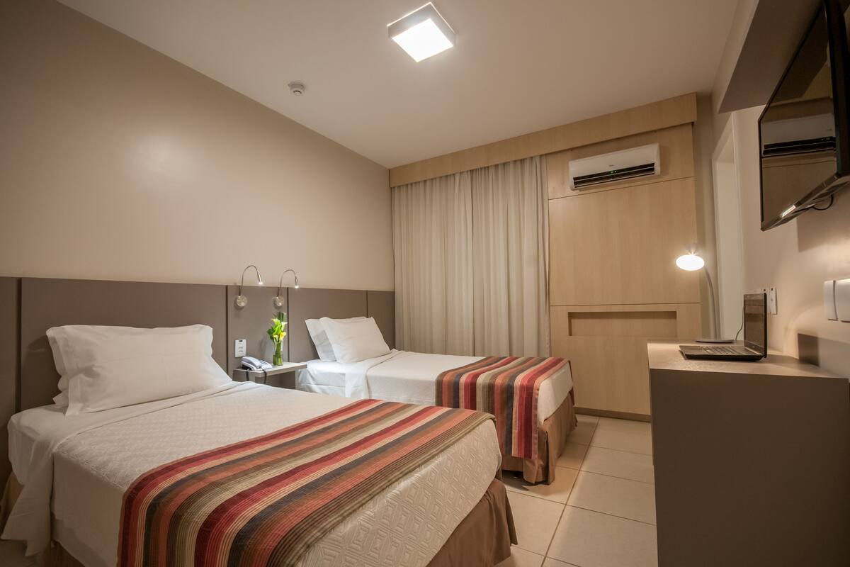 A hotel room with two beds, a desk with a laptop, a mirror, and an air conditioning unit.