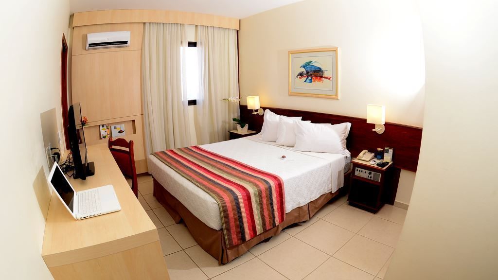 A hotel room with a bed, a desk with a laptop, a telephone, and a nightstand with a lamp.