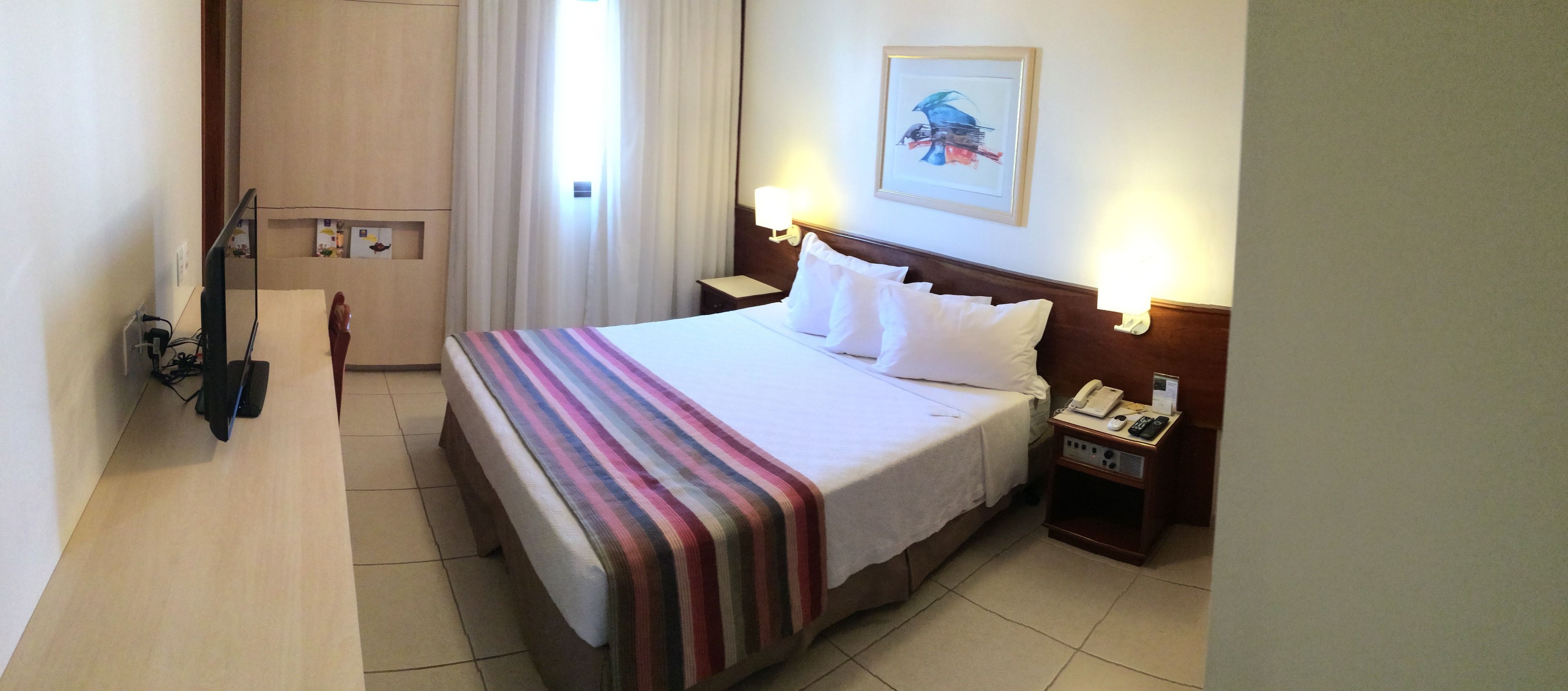 Una camera d'albergo con un letto, un comodino con telefono e lampada, una scrivania con computer e una finestra con tende.