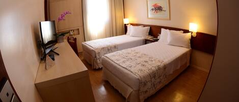 A hotel room with two beds, a desk, a TV, and a window with curtains.