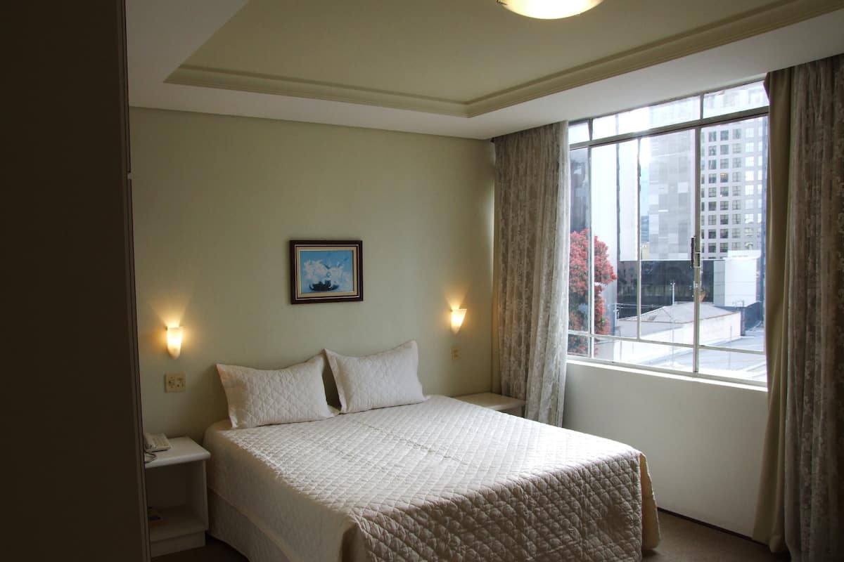 Une chambre d’hôtel avec un lit, une table de chevet, une lampe fixée au mur, une fenêtre avec des rideaux et un tableau encadré accroché au mur.