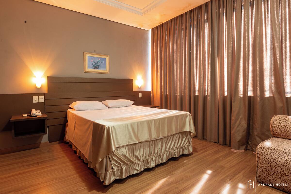Une chambre d’hôtel avec un lit, des lampes de chevet, un téléphone, un tableau au mur et une fenêtre avec des rideaux.