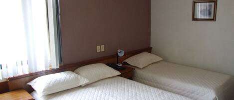 Habitación de hotel con dos camas, una mesita de noche de madera, una pequeña mesa y una ventana con cortinas.