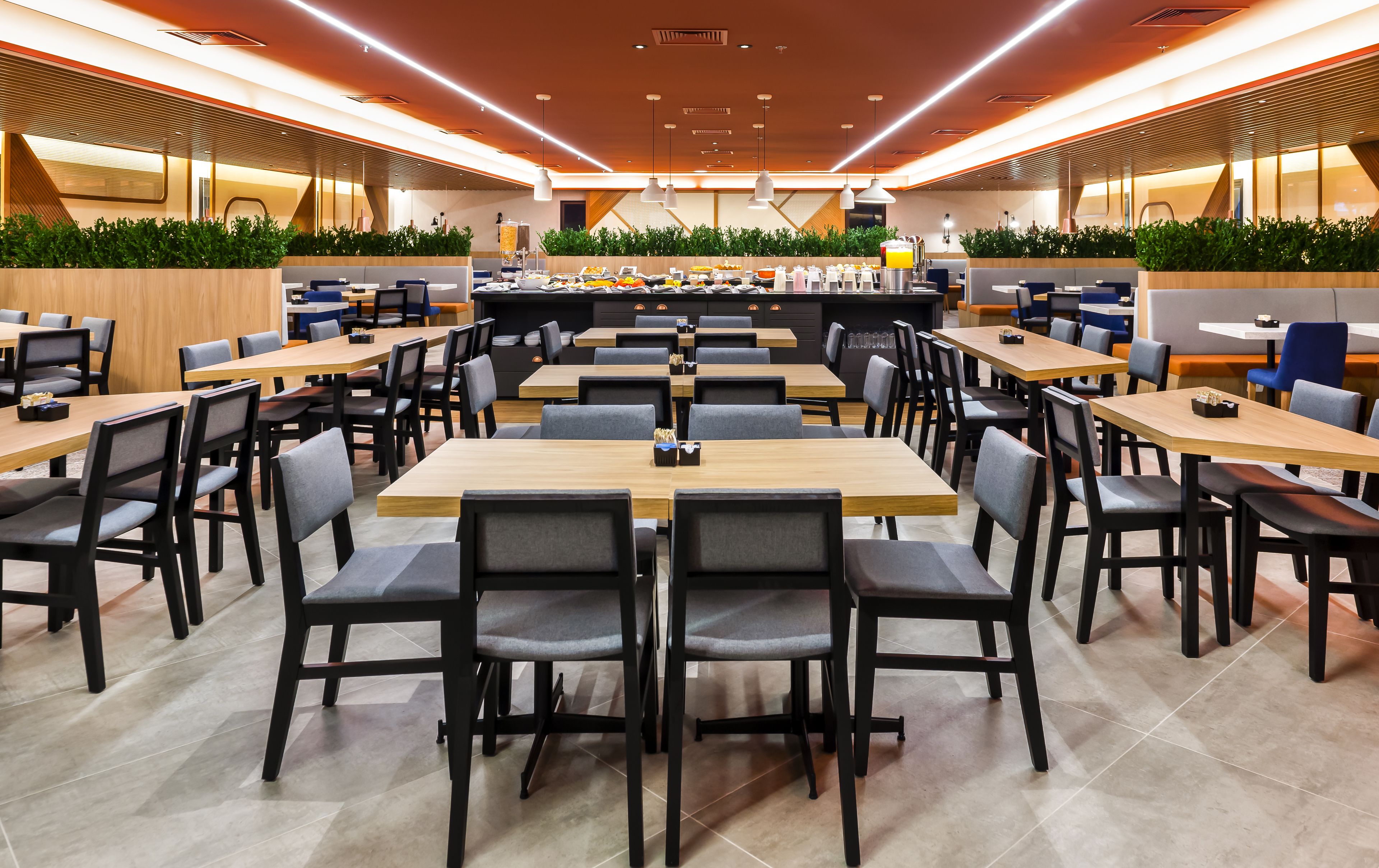 A modern dining area with multiple tables and chairs, a central counter with food and drinks, and green plants along the walls.