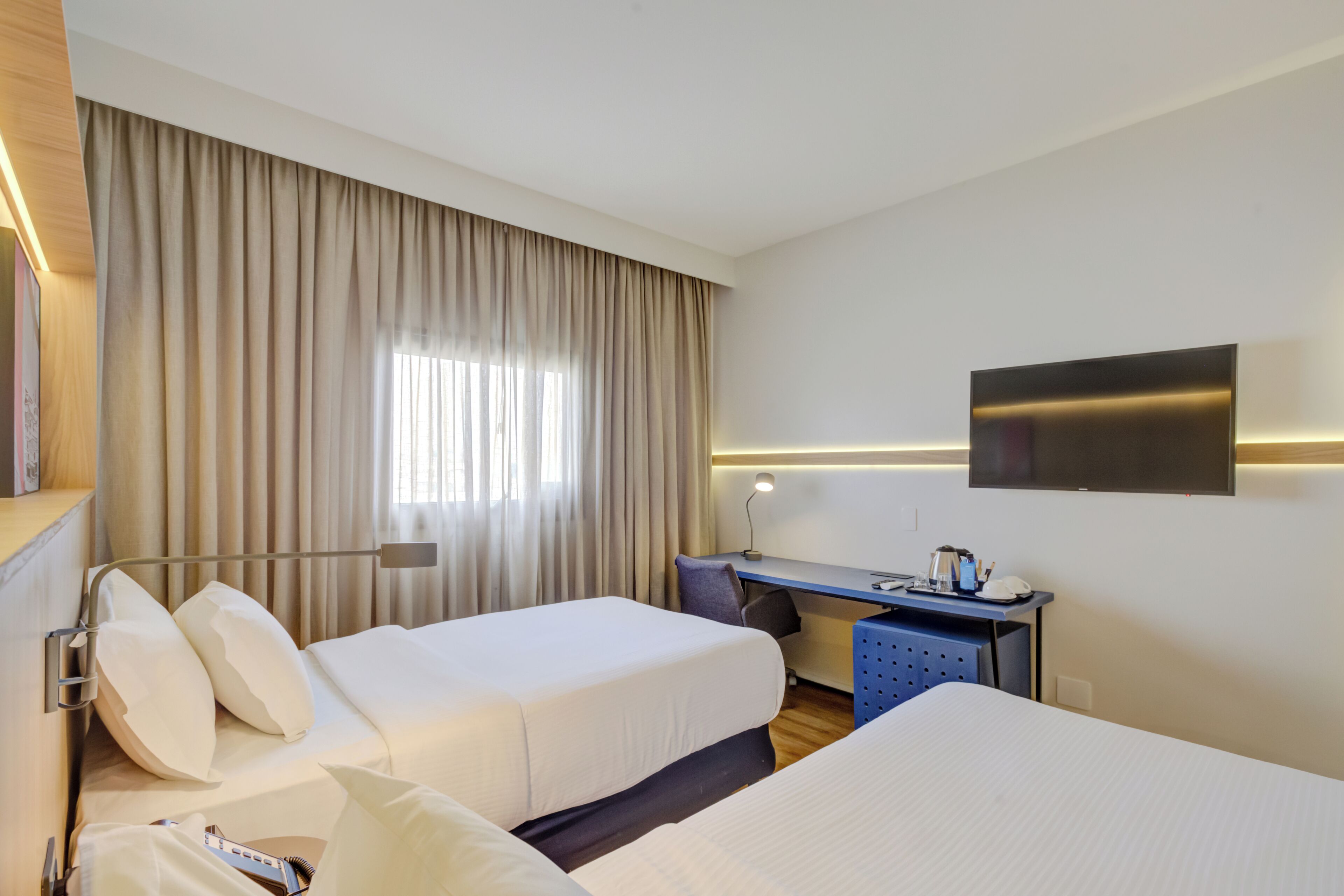 A hotel room with two beds, a desk, a TV, and a window with curtains.