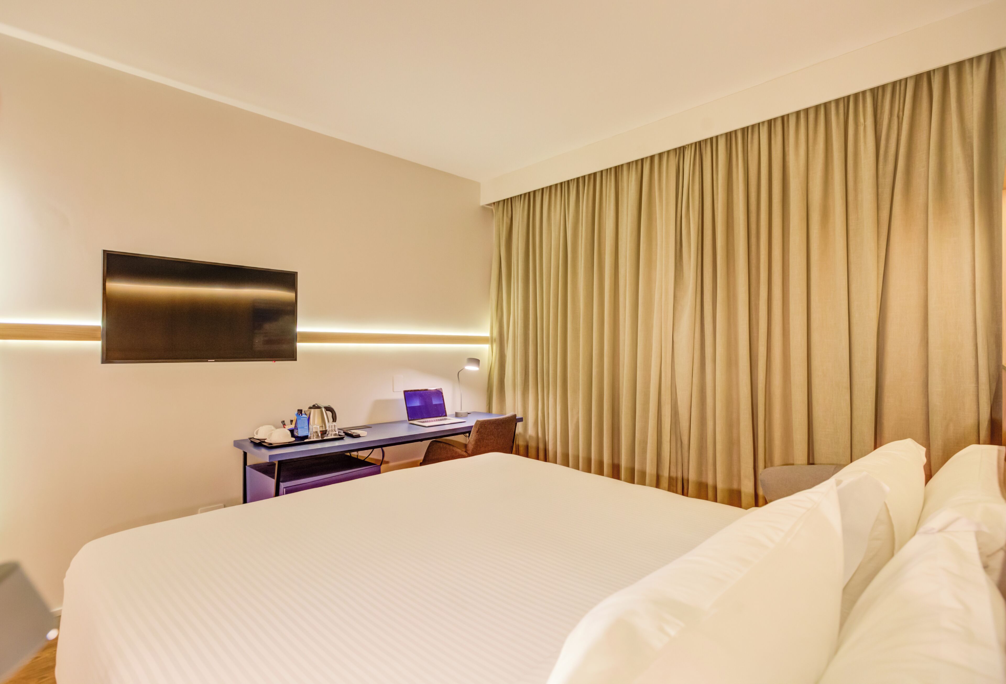 A hotel room with a large bed, a desk with a laptop, a flat-screen TV mounted on the wall, and a curtain-covered window.