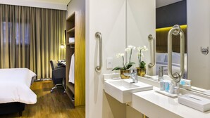Habitació d'hotel amb un llit, una taula de nit, una cadira, un bany amb un lavabo i un mirall, i una finestra amb cortines.