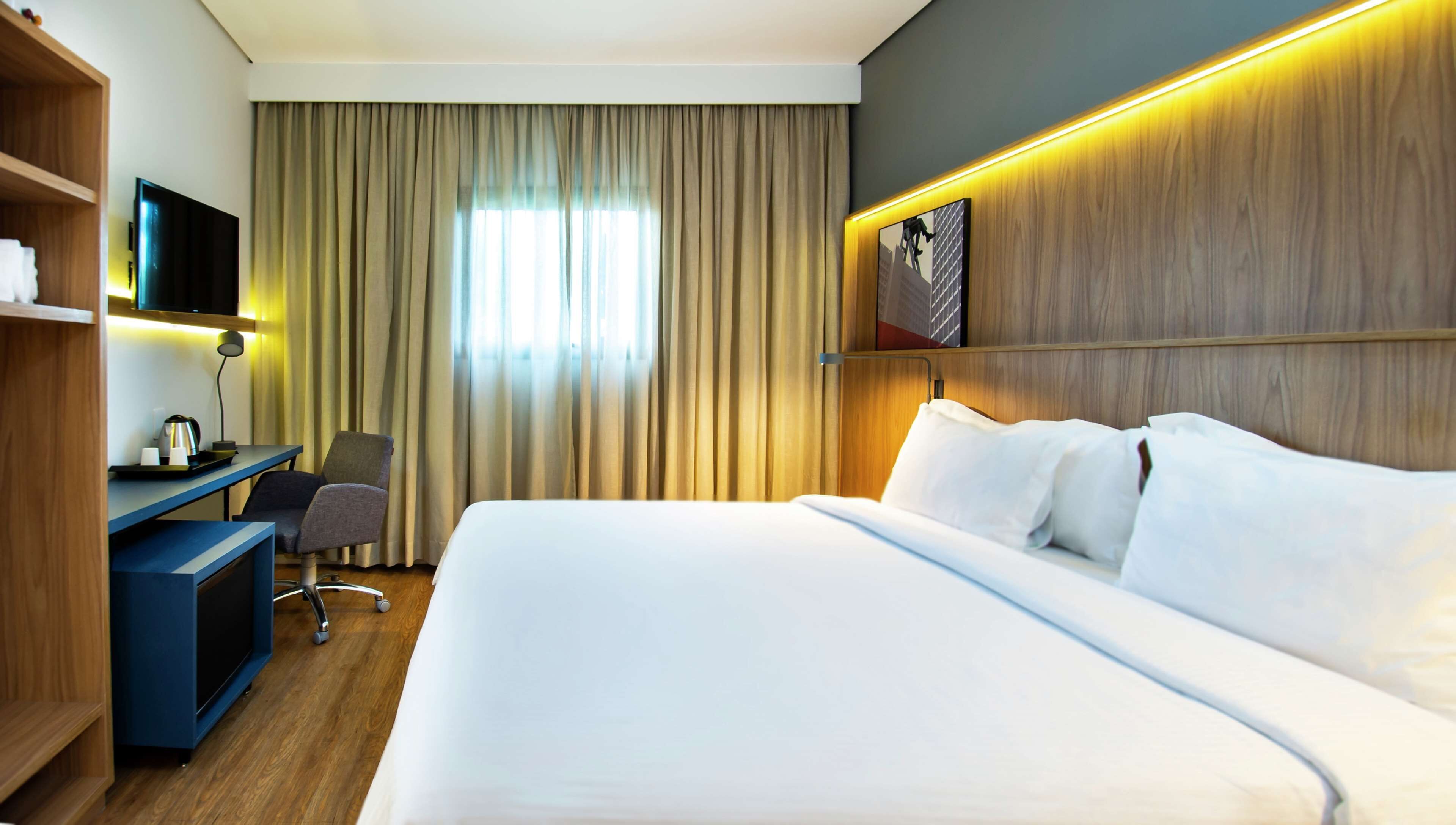 A hotel room with a large bed, a desk with a chair, a television, and a wooden shelf.