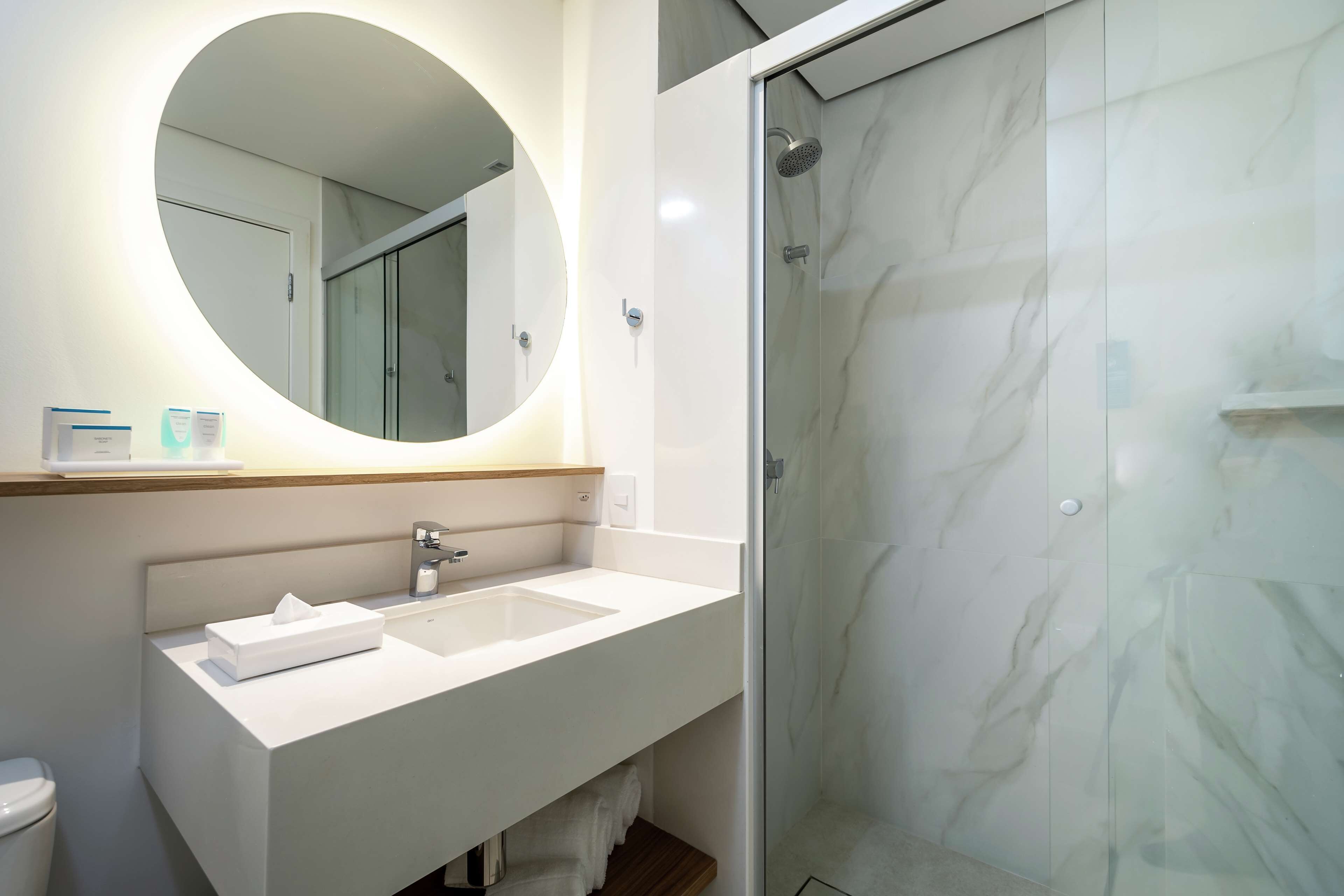 Une salle de bain moderne avec un miroir rond, un lavabo blanc et une douche à parois de verre.