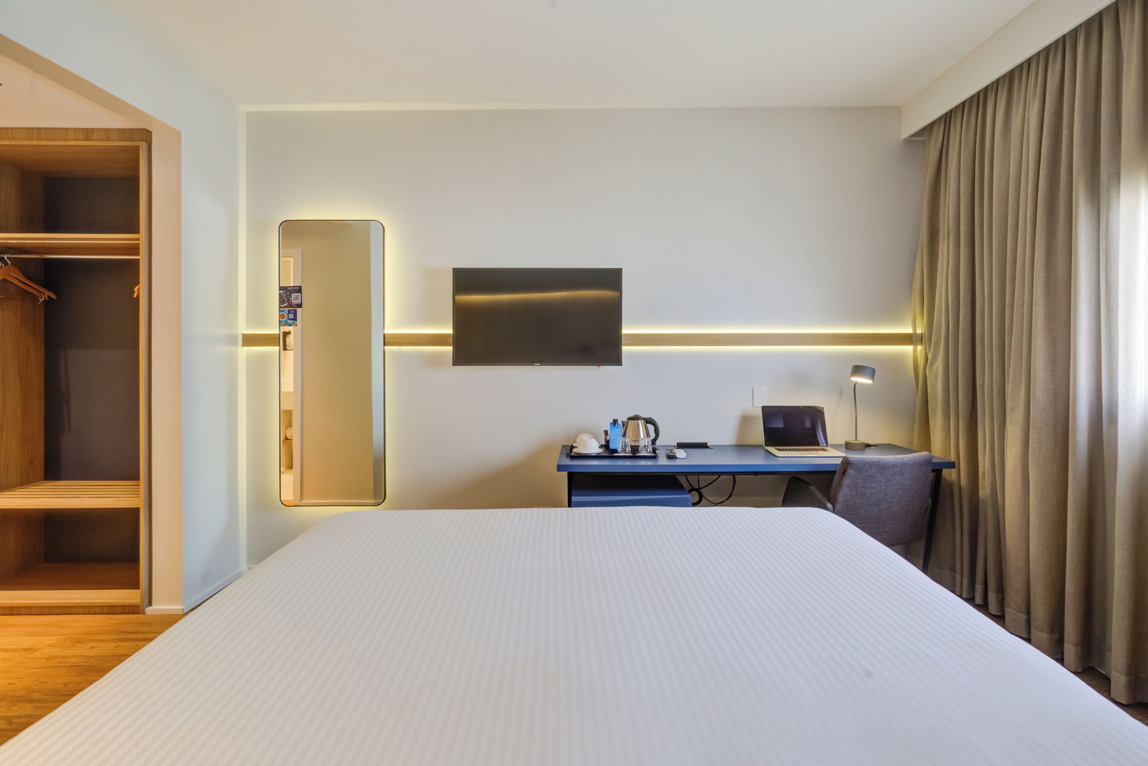 Una camera d'albergo con un letto, una scrivania con un computer, uno specchio e un armadio.