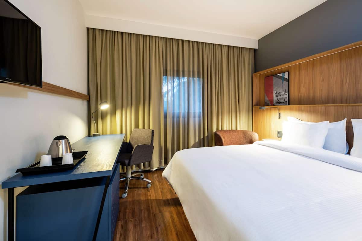 Ein Hotelzimmer mit einem Bett, einem Schreibtisch mit Lampe, einem Sessel und einem Fernseher an der Wand.