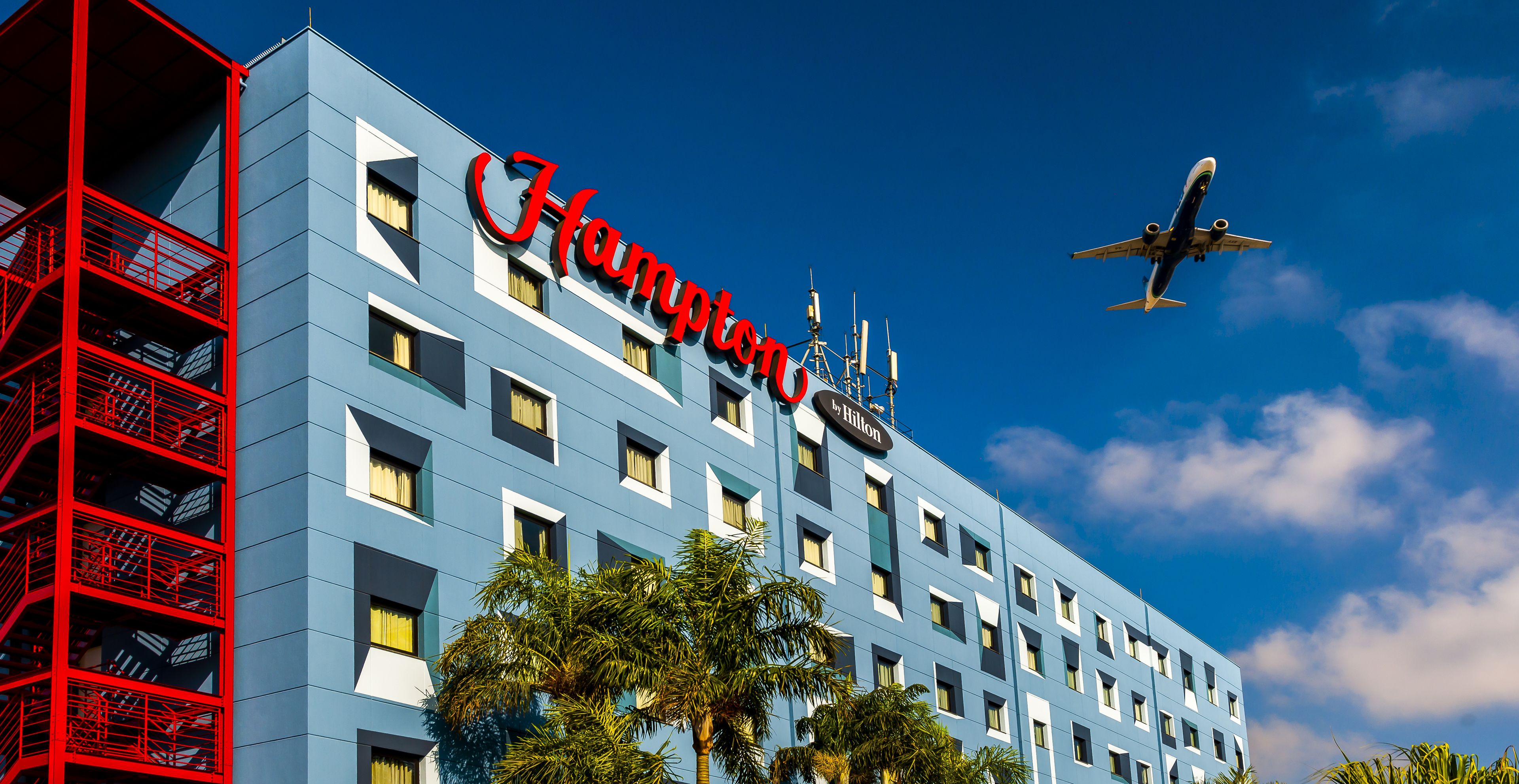 Un hôtel Hampton by Hilton, avec un ciel bleu azur et un avion en arrière-plan.
