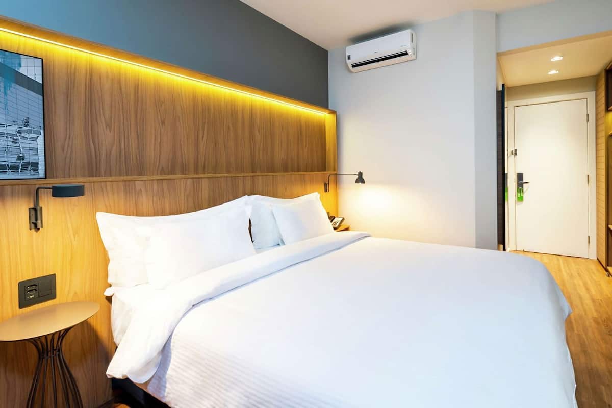 Ein Hotelzimmer mit einem hölzernen Kopfteil, einem Bett mit weißer Bettwäsche, einem Nachttisch, einer Klimaanlage und einem Schrank mit Tür.