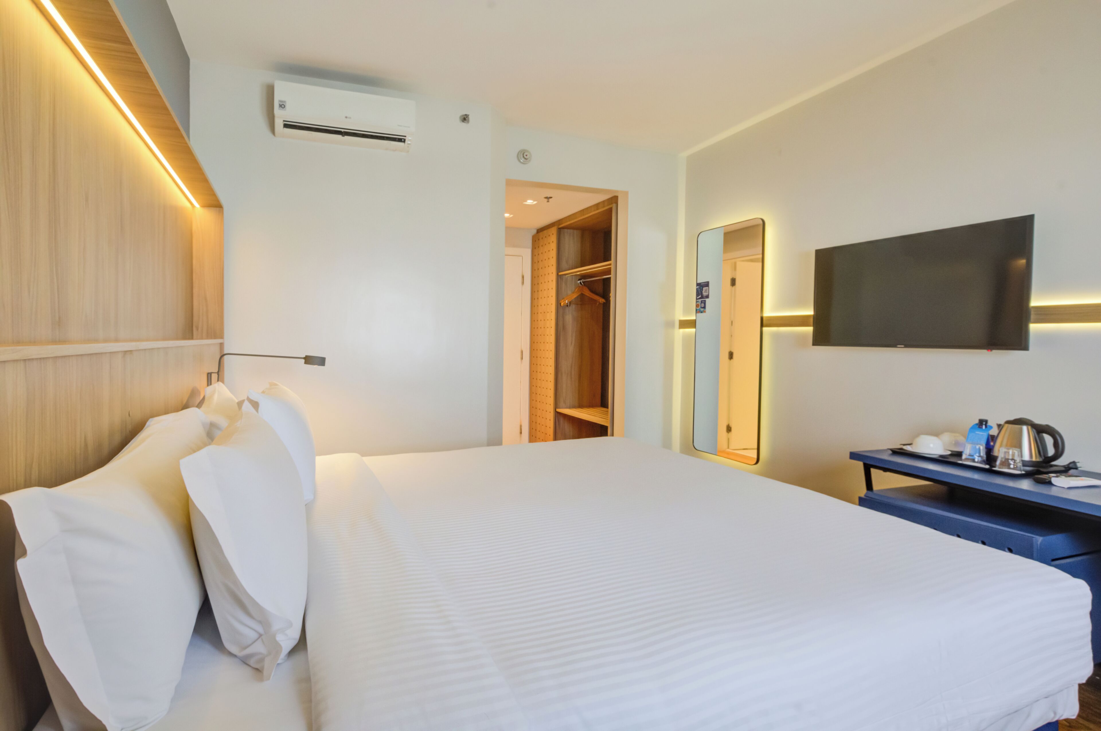 A hotel room with a bed, a wardrobe, a desk with a kettle, and a flat-screen TV.