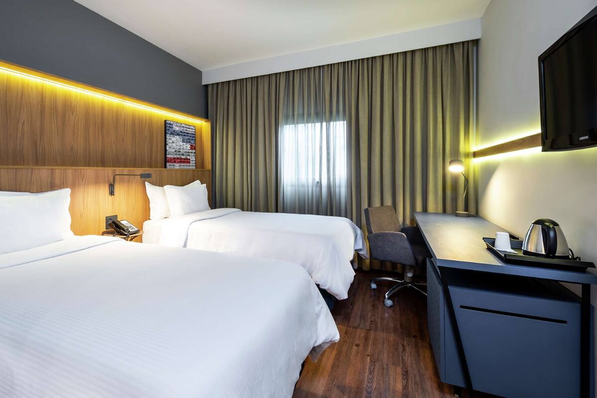 Ein Hotelzimmer mit zwei Betten, einem Schreibtisch mit Stuhl, einer Schreibtischlampe, einem Telefon und einem an der Wand montierten Fernseher.