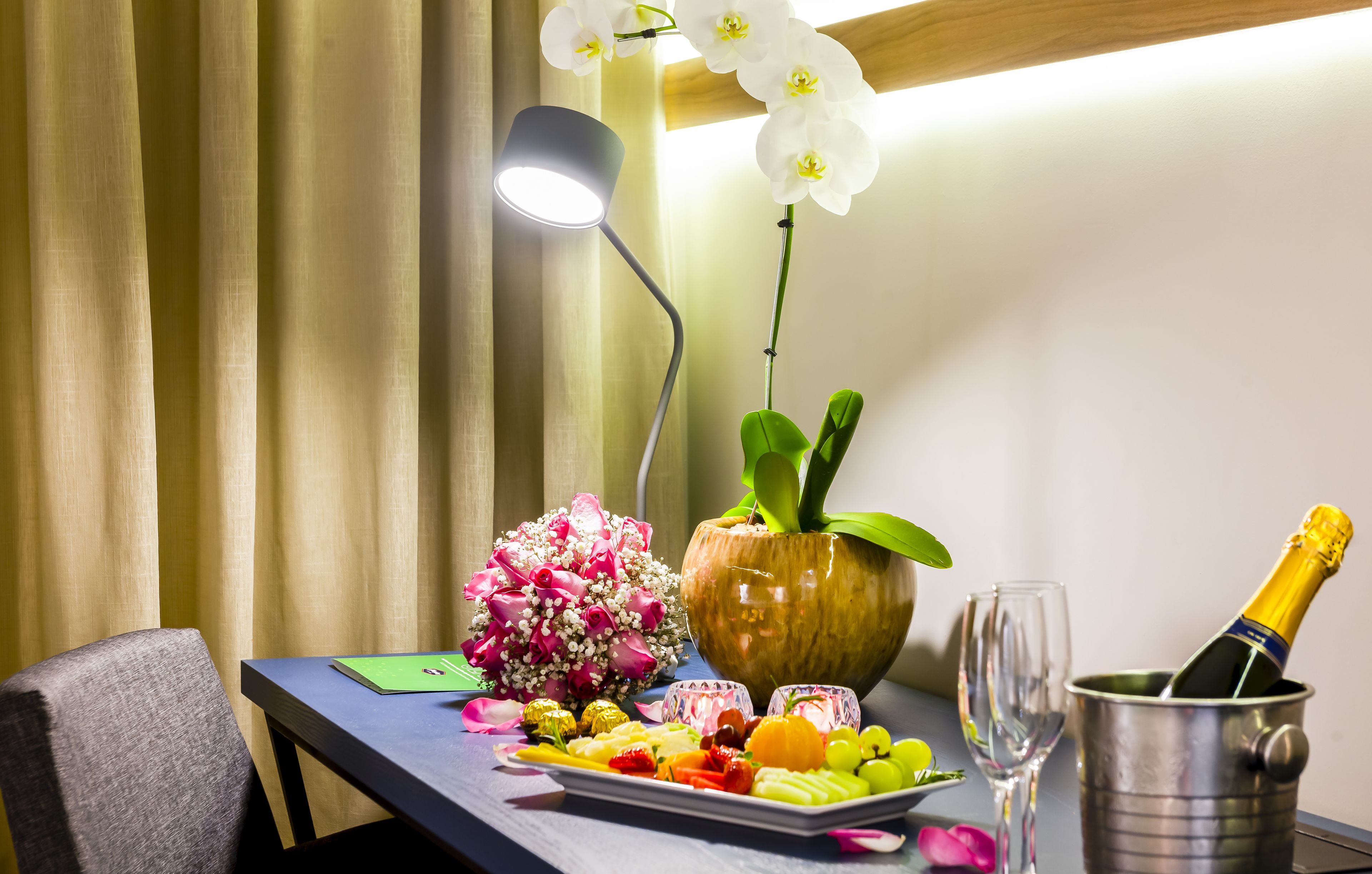 Una mesa con una botella de champán, una copa, una bandeja de frutas, un jarrón con flores y una lámpara.