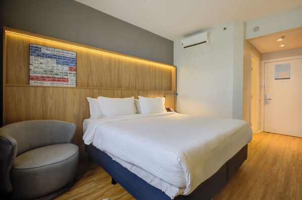 A hotel room with a large bed, a chair, a wooden headboard, a wall-mounted air conditioner, and a door.