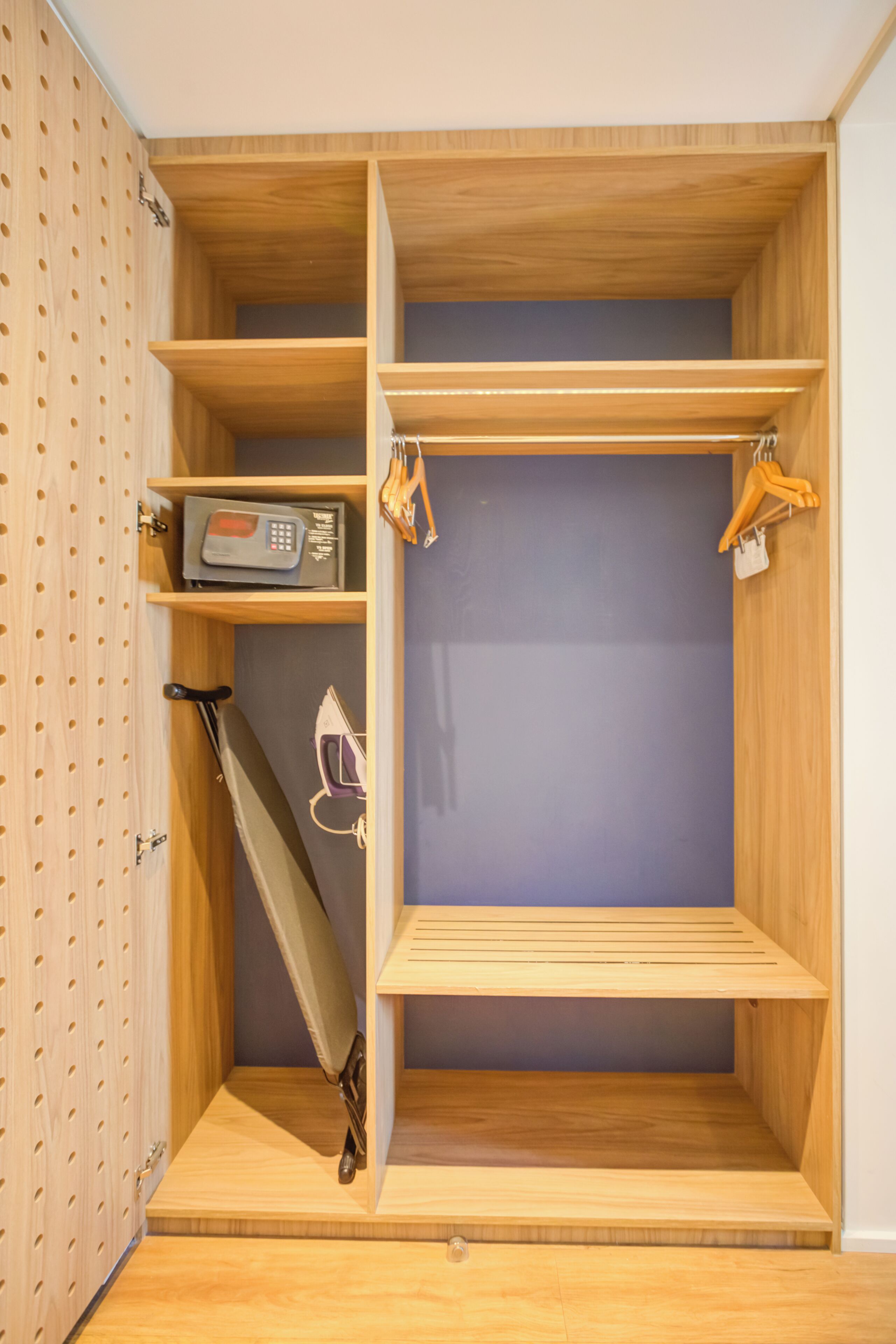 Un placard en bois avec des étagères et une tringle pour suspendre les vêtements, une petite planche à repasser et une radio.