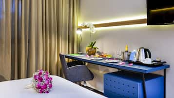 A hotel room with a neatly arranged desk, a vase with an orchid, a bottle of champagne, a coffee maker, and a TV mounted on the wall.