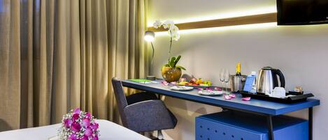 Habitación de hotel con un escritorio ordenado, un florero con una orquídea, una botella de champán, una cafetera y un televisor montado en la pared.