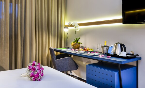 A hotel room with a neatly arranged desk, a vase with an orchid, a bottle of champagne, a coffee maker, and a TV mounted on the wall.