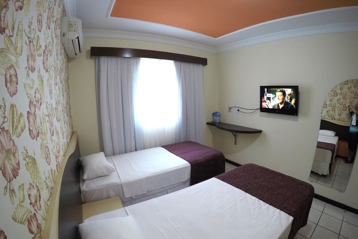A hotel room with two beds, a television, a window with curtains, and a floral wallpaper.
