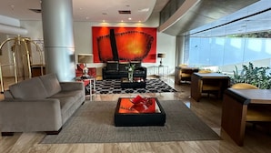 A spacious lobby with a modern design, featuring a large seating area with sofas and a coffee table, a large abstract painting on the wall, and a view of the outdoors through large windows. - Hotel Saint Paul (São José do Rio Preto)