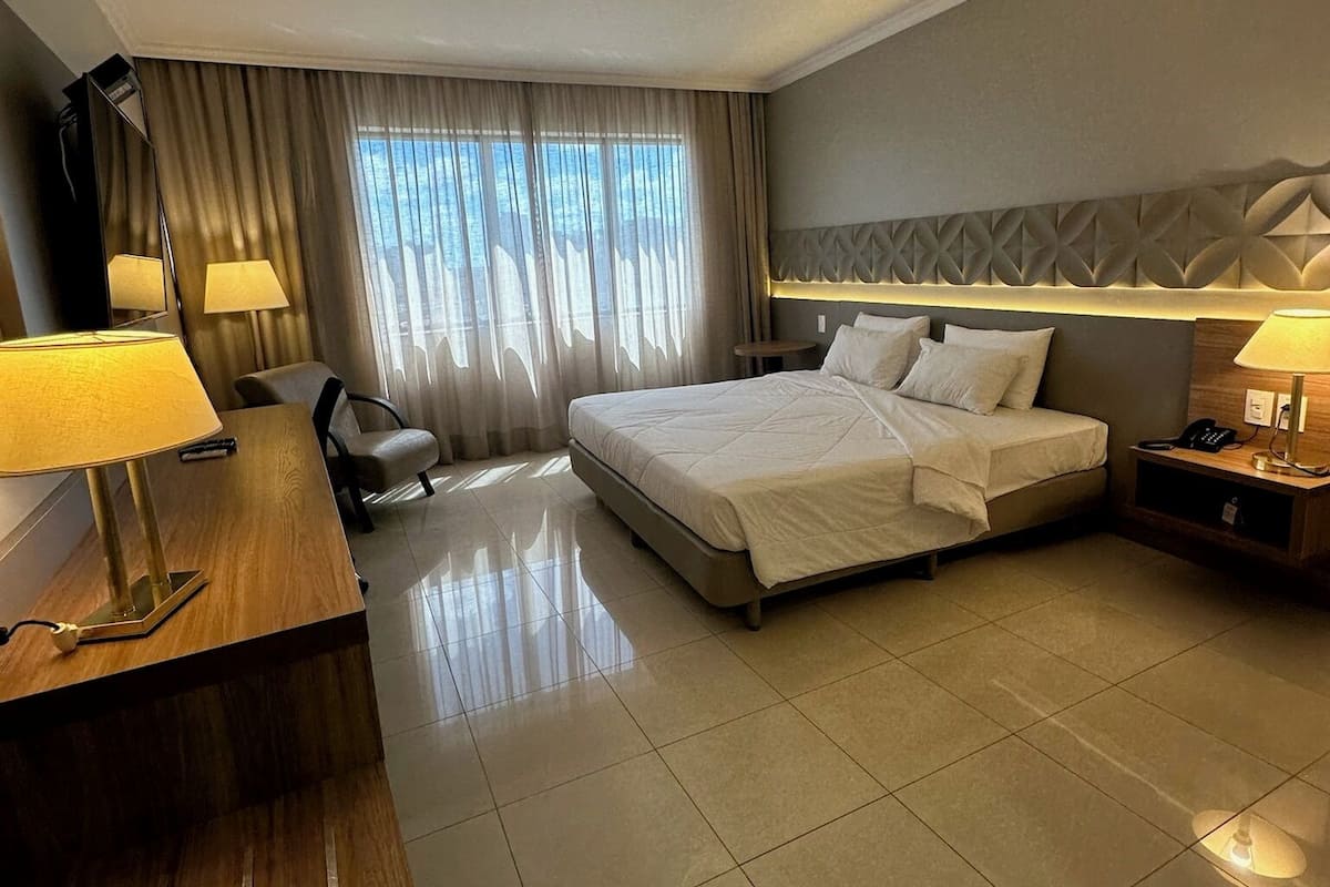 A hotel room with a large bed, a desk with a lamp, a chair, a bedside table with a lamp, and a window with curtains.