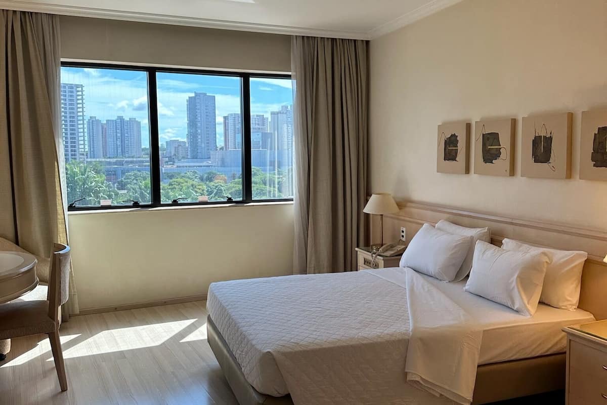 A hotel room with a large bed, a desk, a chair, and a view of the cityscape through the window.