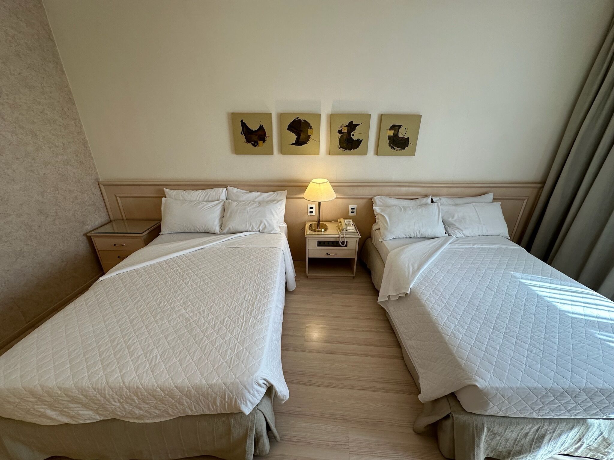 A hotel room with two beds, a nightstand with a lamp, and three wall art pieces.