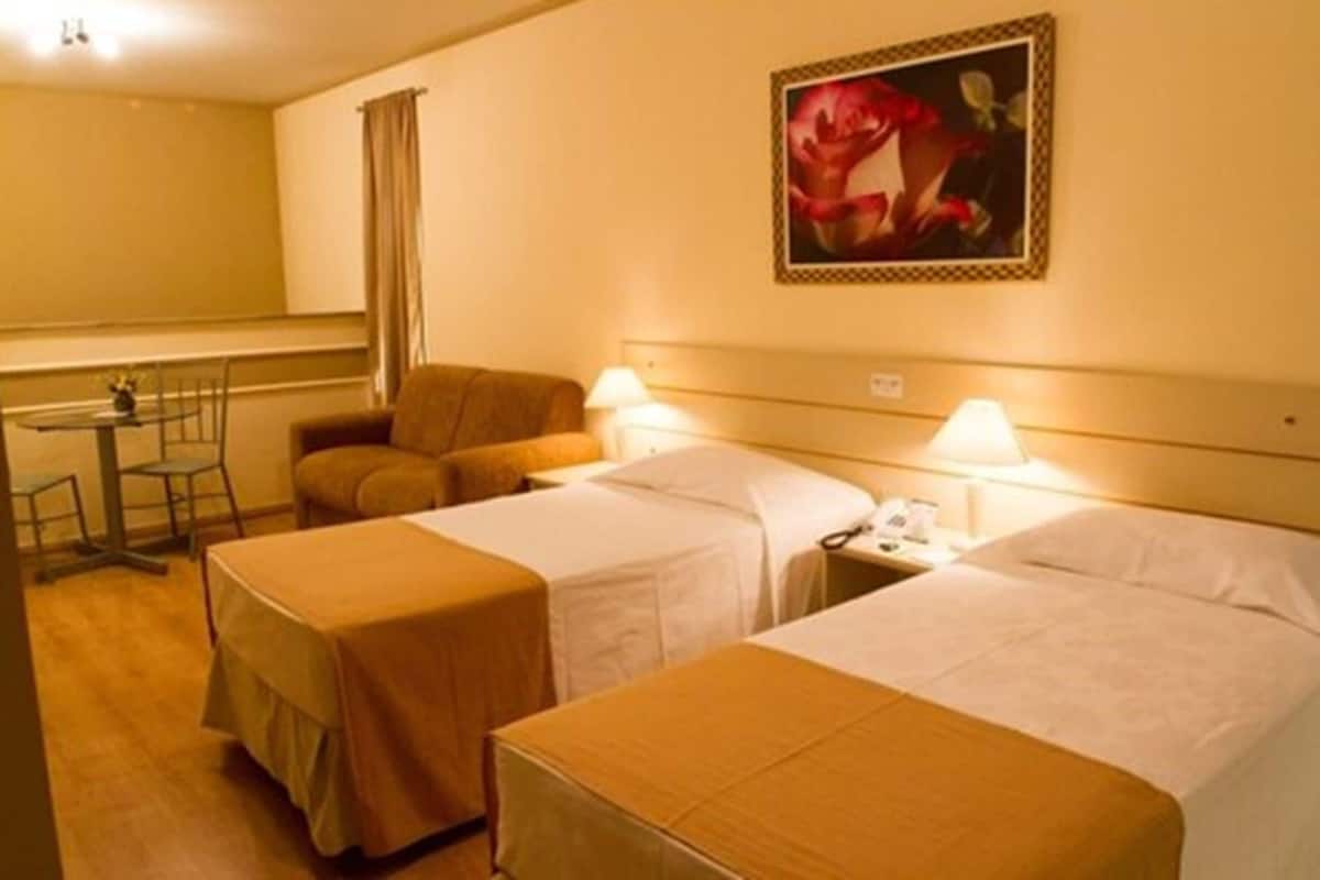 A hotel room with two beds, a small table, a chair, and a painting on the wall.