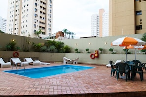 A swimming pool with lounge chairs and a table with an umbrella, surrounded by a brick patio and a low wall with plants.