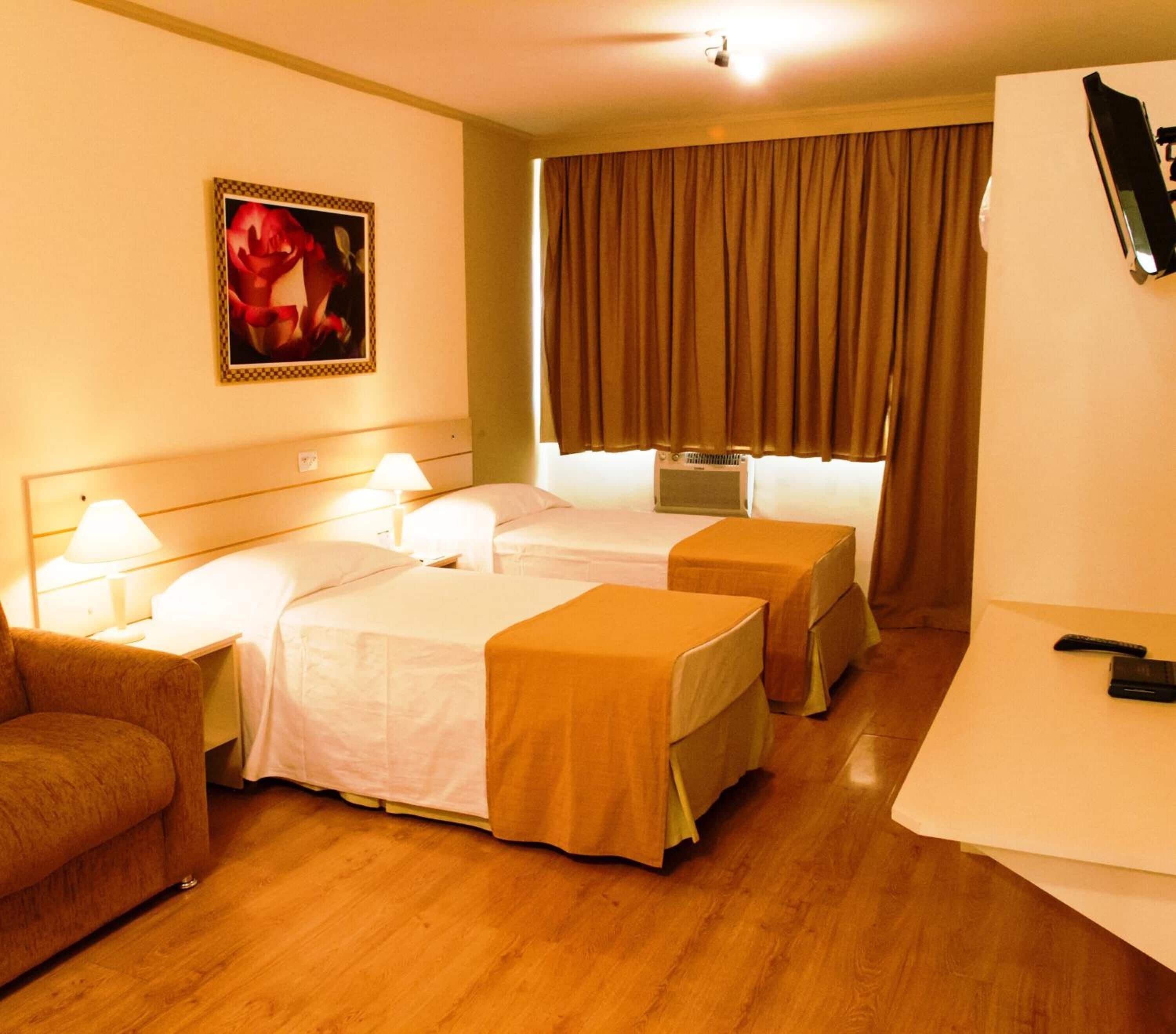 A hotel room with two beds, a desk, a chair, a TV, and a painting on the wall.