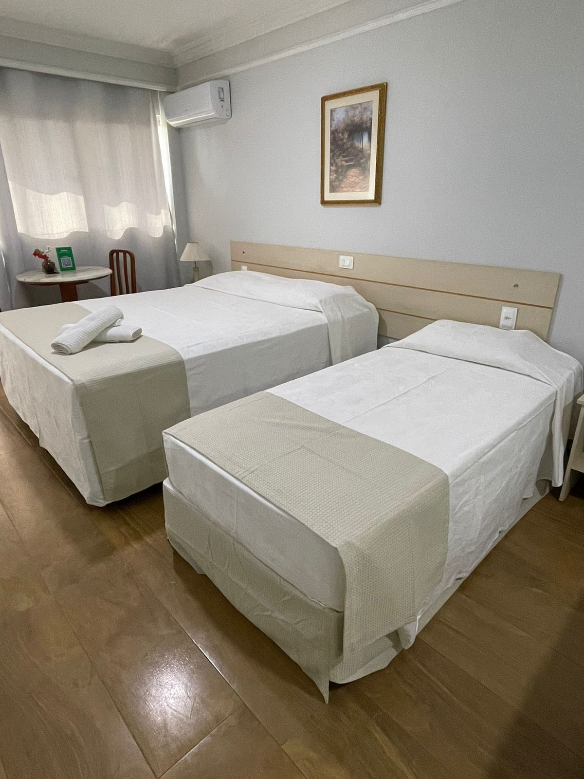 A hotel room with two beds, a small table with a chair, a lamp, and a framed picture on the wall.