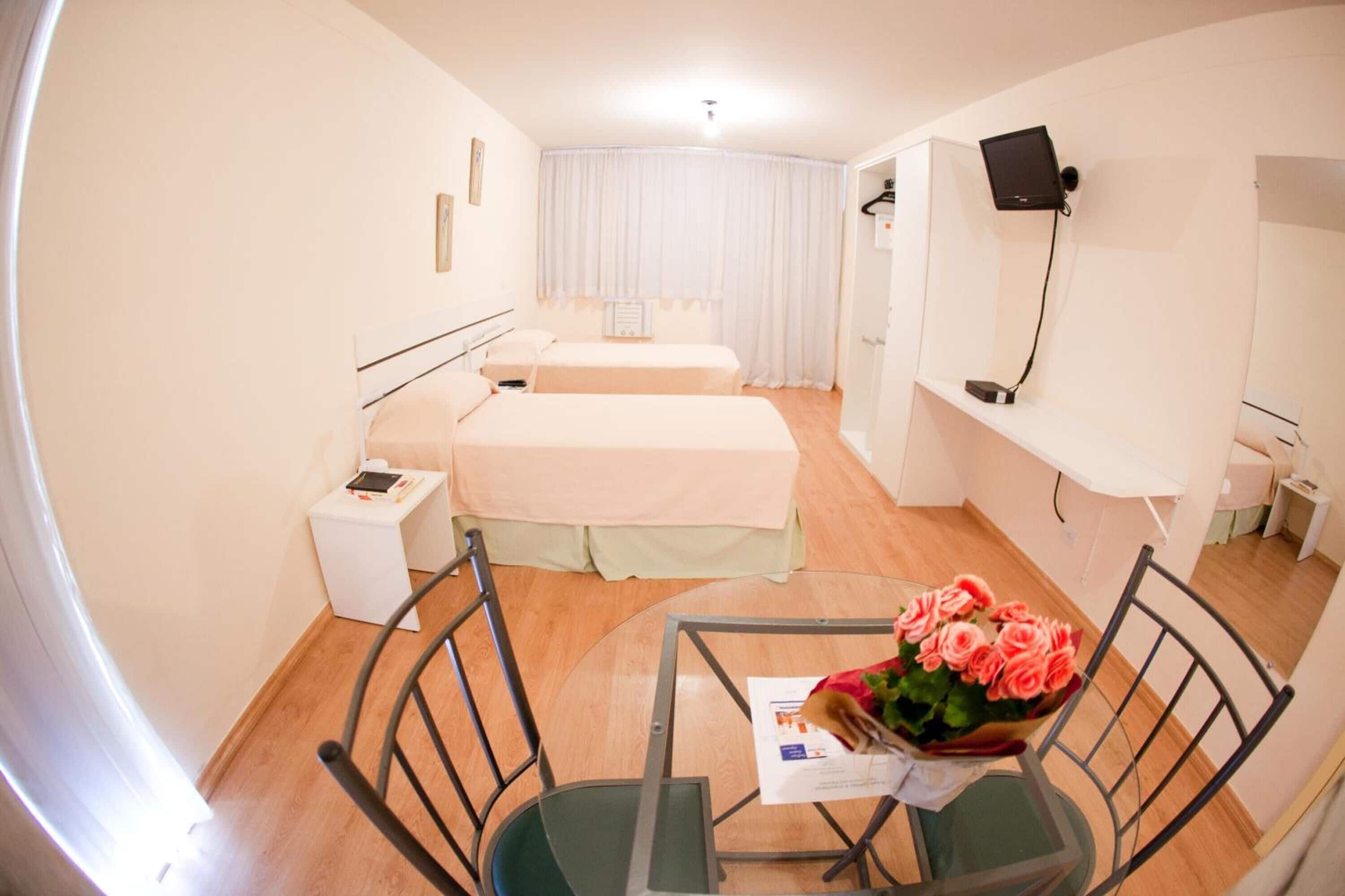 Habitación de hotel con dos camas, una mesita pequeña con un ramo de flores, un televisor montado en la pared y una mesita de noche con una lámpara.