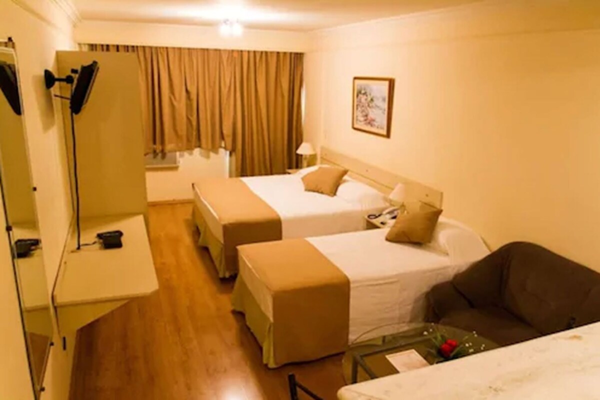 Quarto de hotel com duas camas, um sofá, uma escrivaninha com abajur e um espelho.