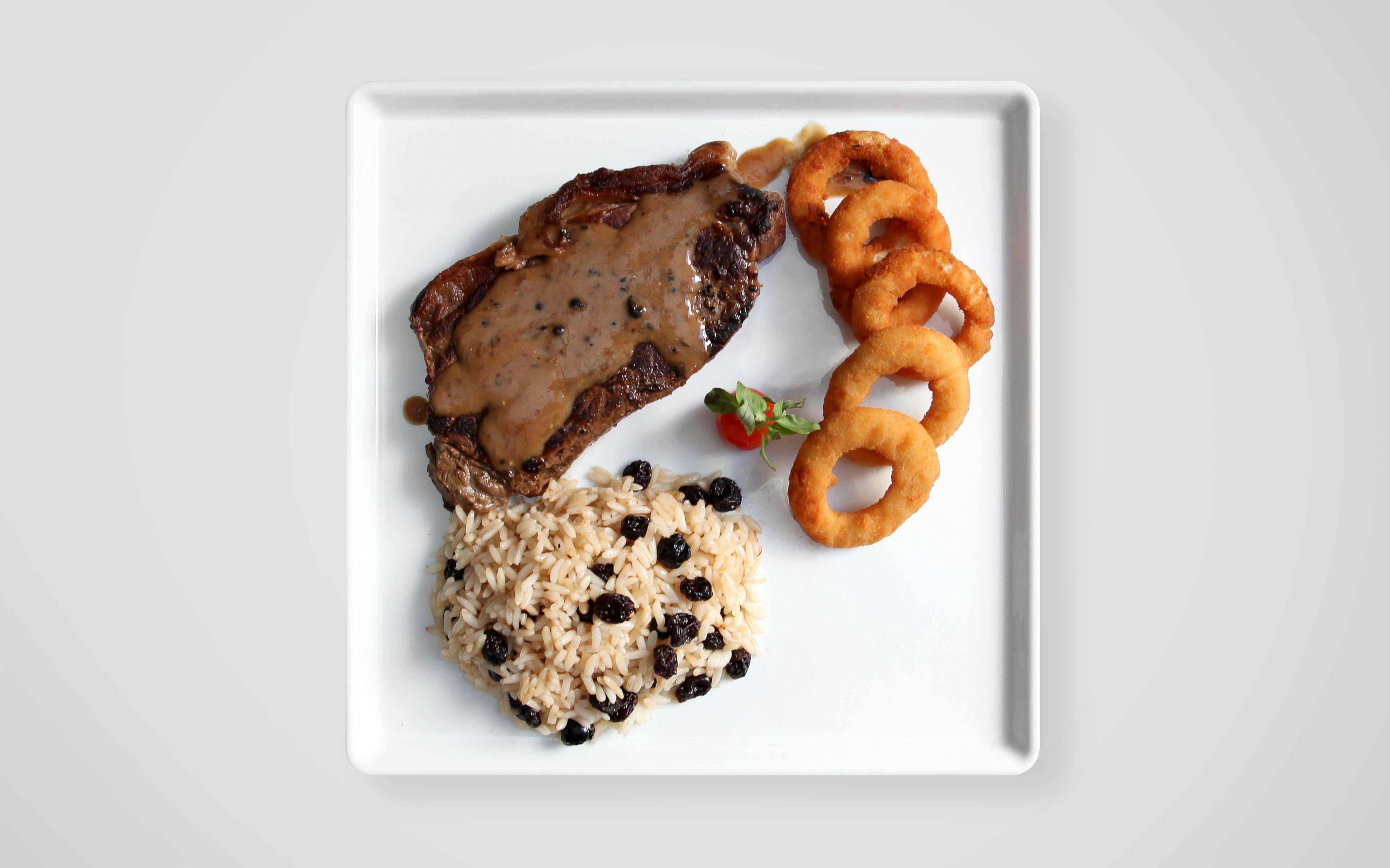 A square plate with a piece of meat topped with gravy, a serving of rice with raisins, and onion rings.