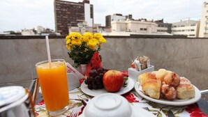 Una mesa con un vaso de jugo de naranja, un ramo de flores amarillas, un plato con uvas, una manzana y pasteles sobre un mantel floral, con vista a un paisaje urbano de fondo.