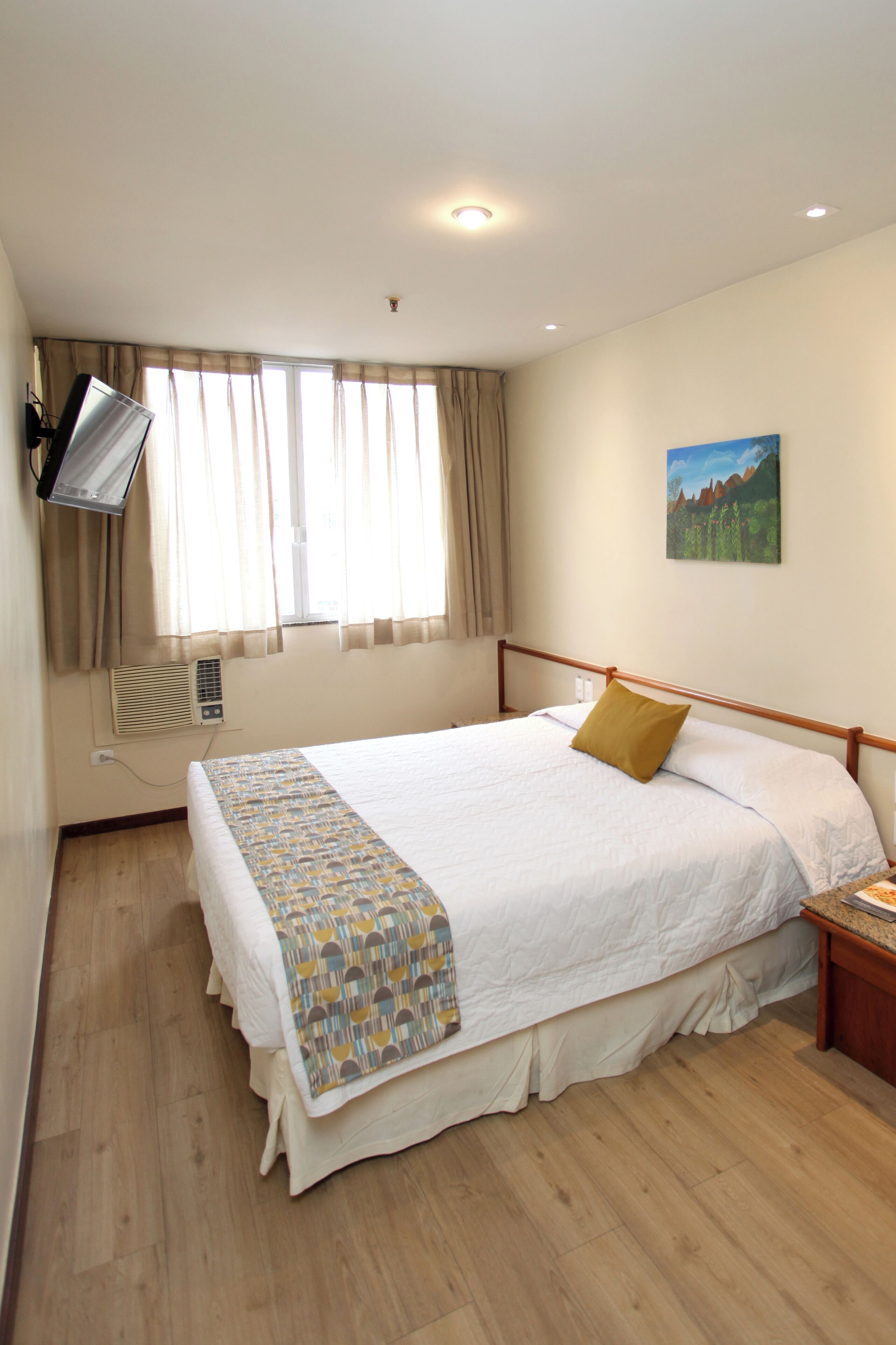 Ein Hotelzimmer mit einem großen Bett, einem flachbildfernseher an der Wand, einem Fenster mit Vorhängen und einem Gemälde.