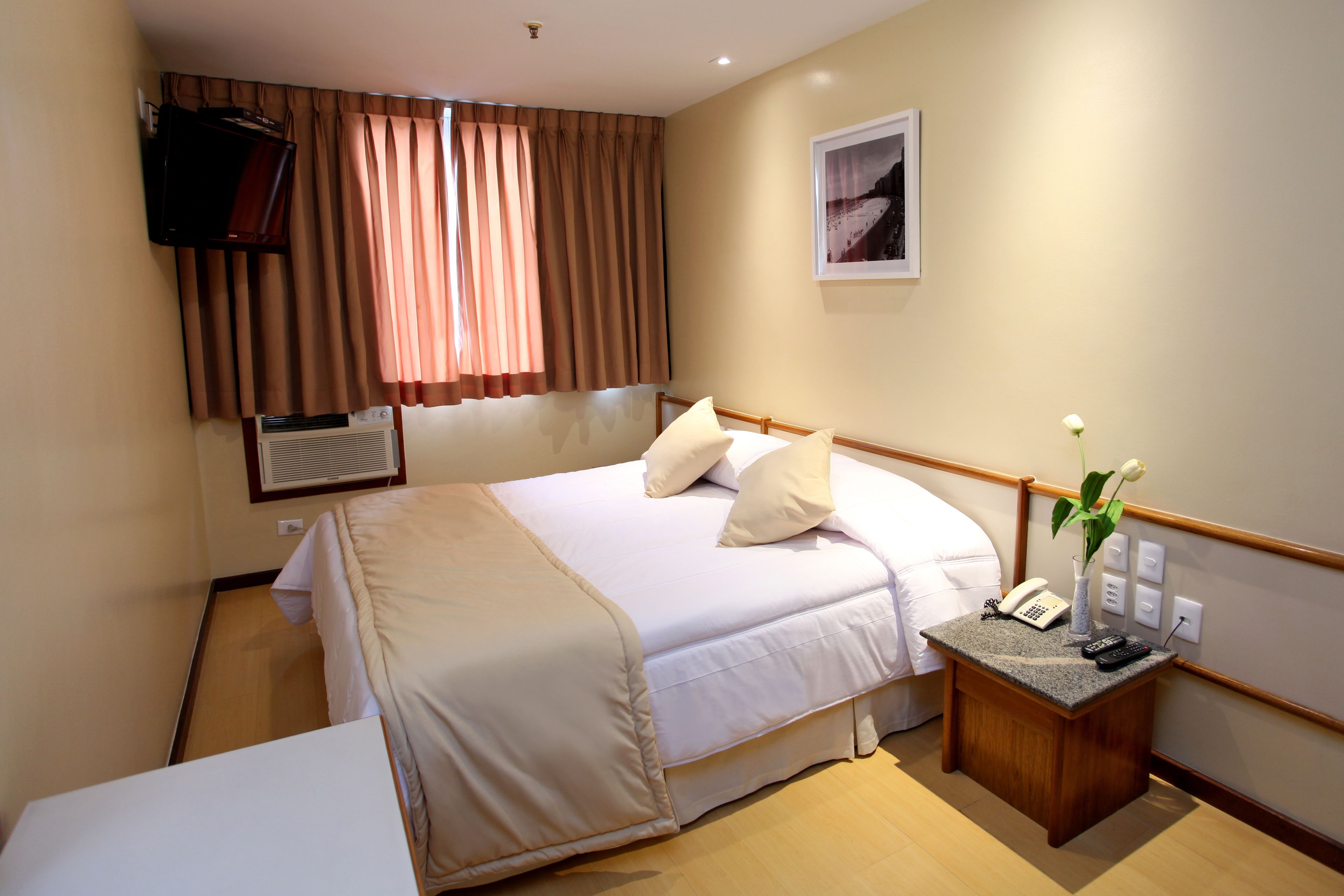 A hotel room with a bed, bedside table with a phone and remote, a TV mounted on the wall, and a window with curtains.