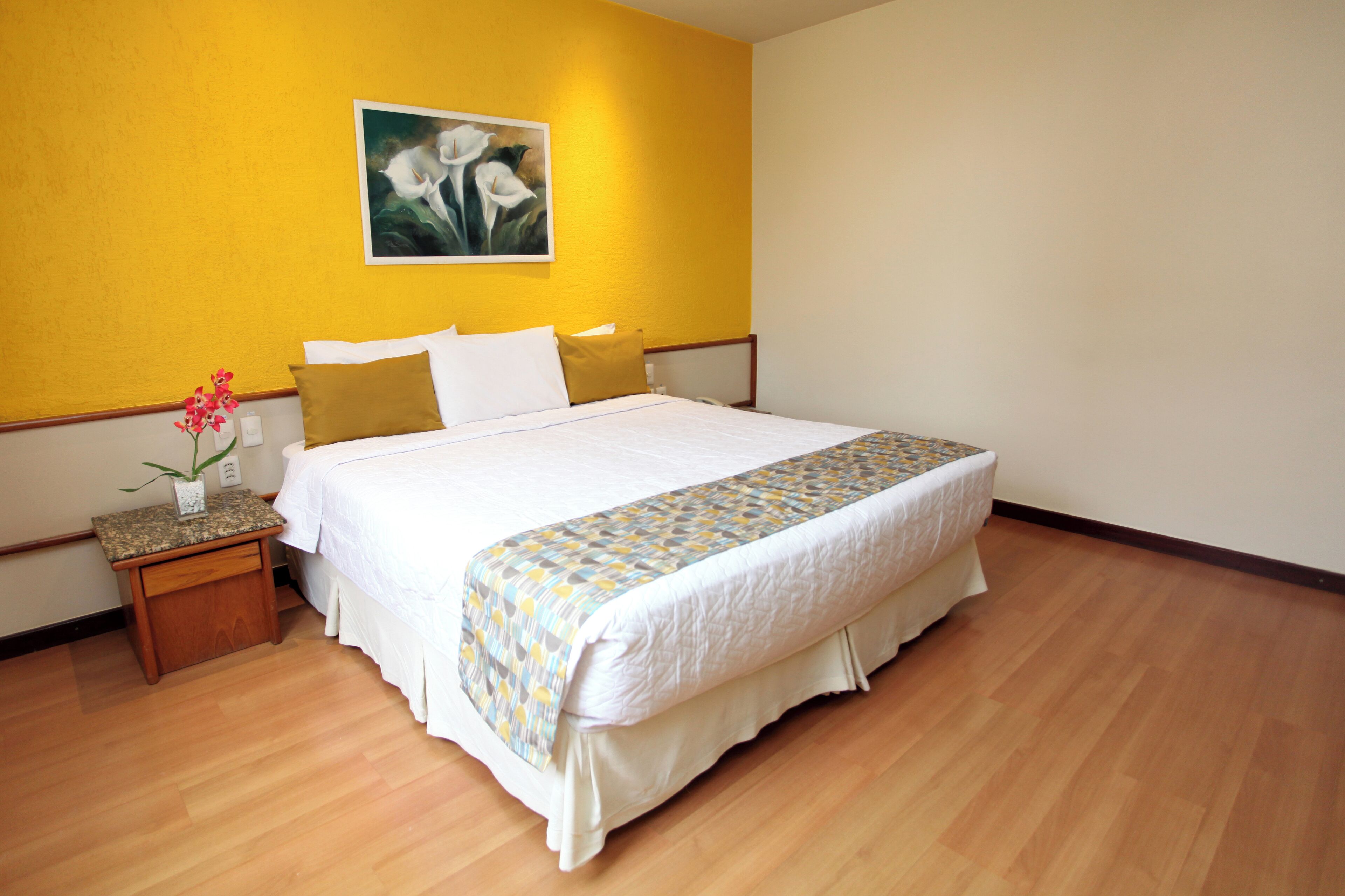 A hotel room with a bed, a nightstand, a vase with flowers, and a painting on the wall.