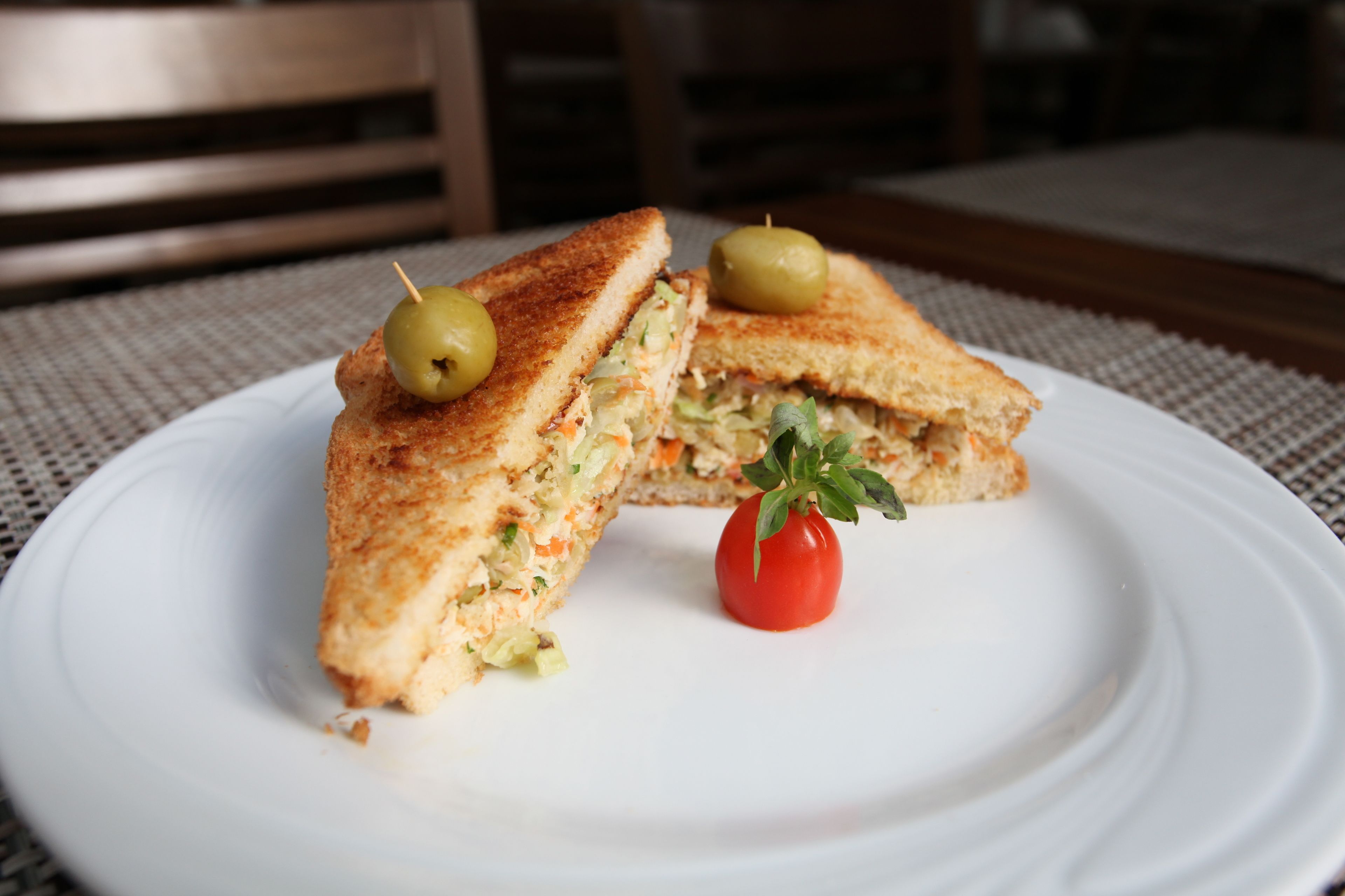 Un sandwich sur une assiette blanche, accompagné d’une tomate cerise et de deux olives.