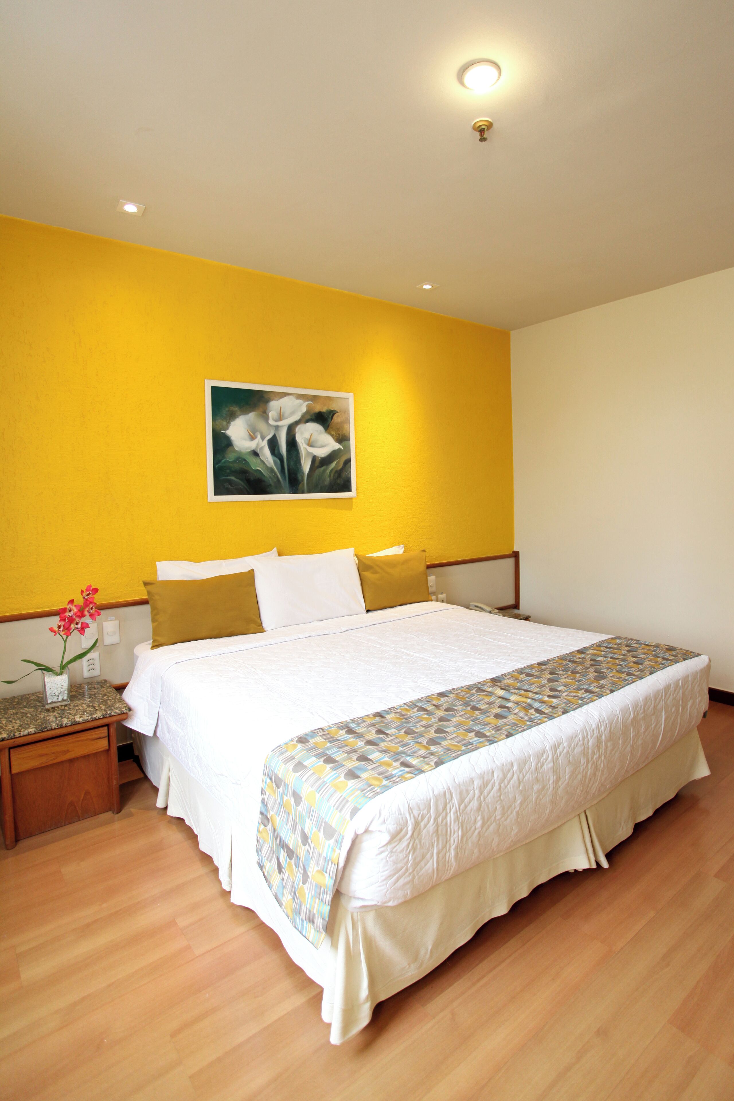 A hotel room with a bed, a nightstand, a vase with flowers, and a painting on the wall.