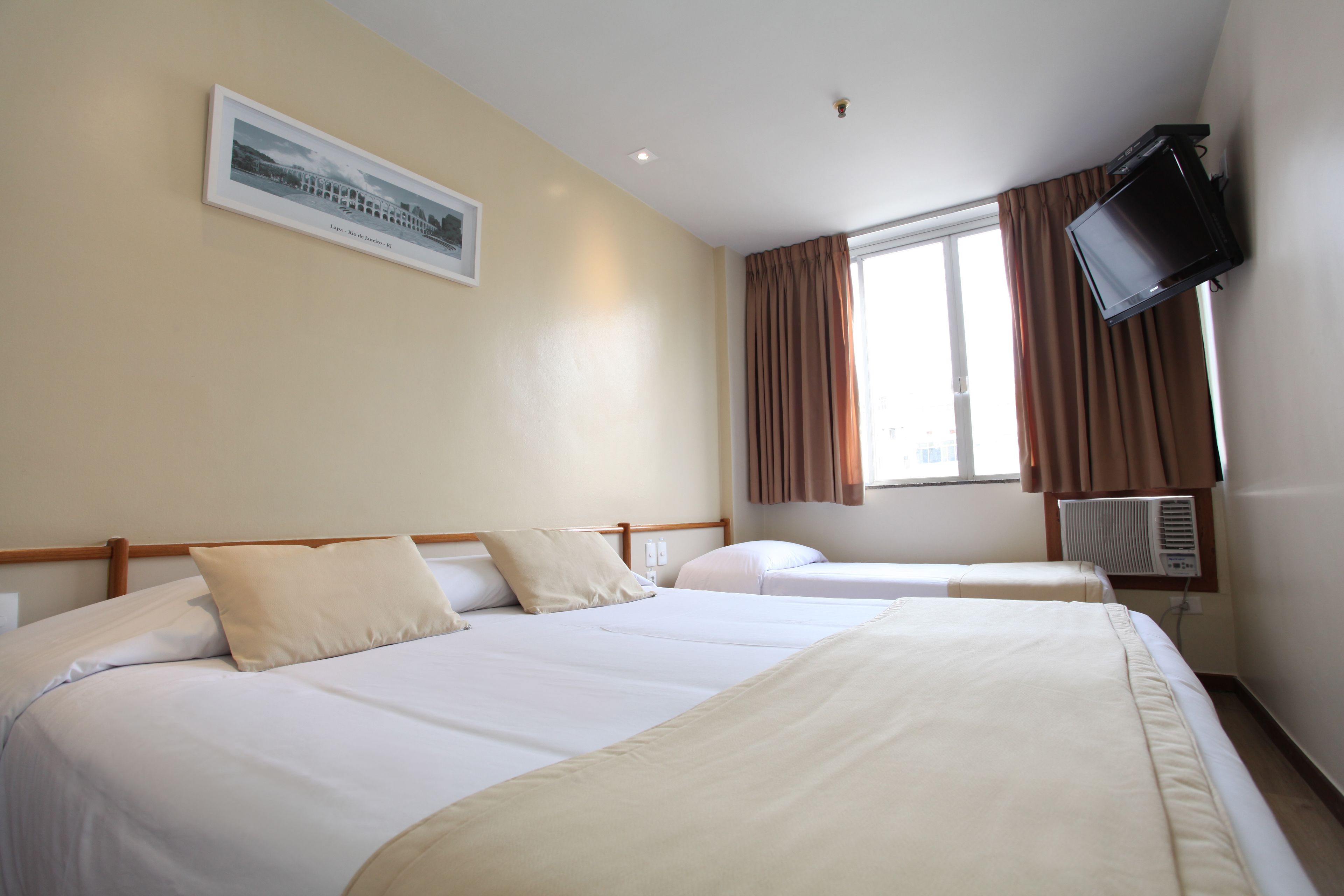 A hotel room with two beds, a television mounted on the wall, and a window with curtains.