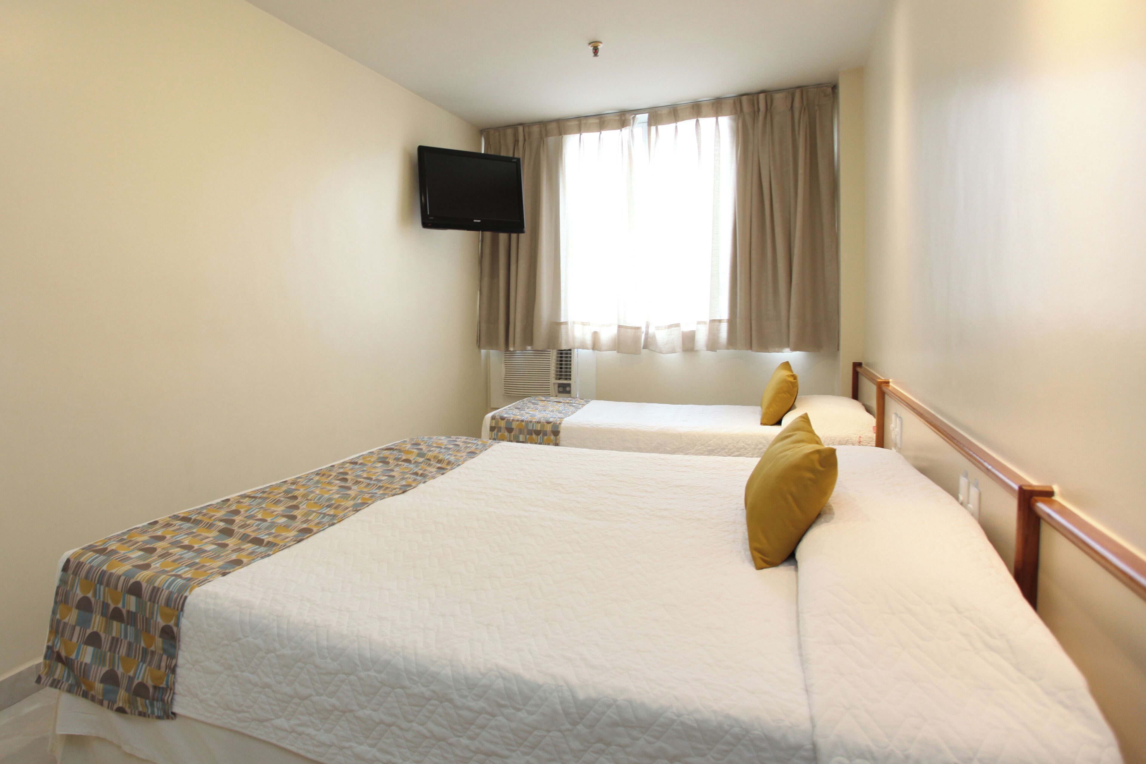 Een hotelkamer met twee bedden, een aan de muur bevestigde televisie en een raam met gordijnen.